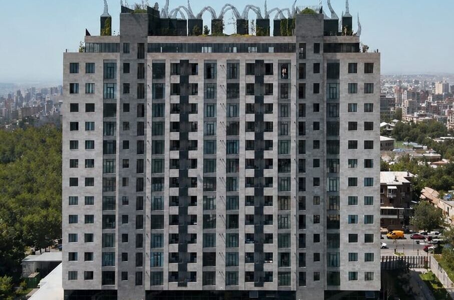 Tall gray high-rise apartment building with decorative loop structures on the roof, overlooking a city park and skyline.