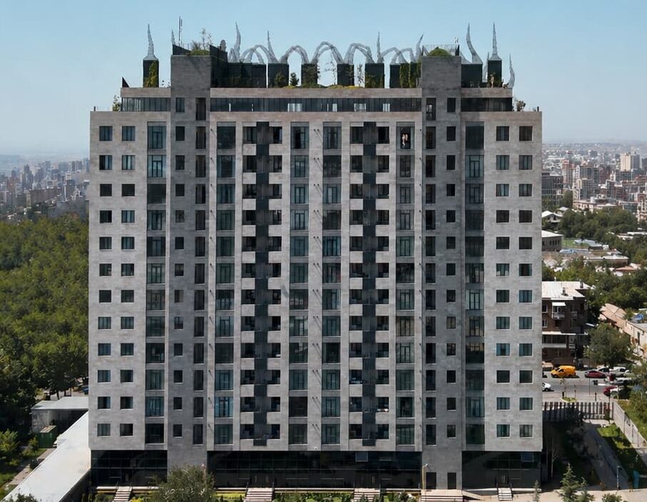 Tall gray high-rise apartment building with decorative loop structures on the roof, overlooking a city park and skyline.