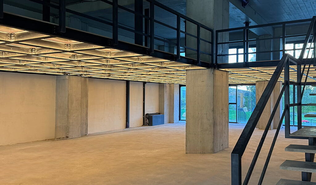 Empty industrial interior with concrete pillars and a mezzanine level, metal staircase on the right side.