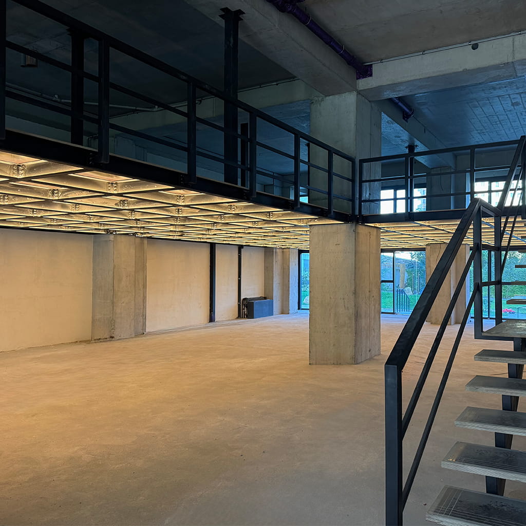 Empty industrial interior with concrete pillars and a mezzanine level, metal staircase on the right side.