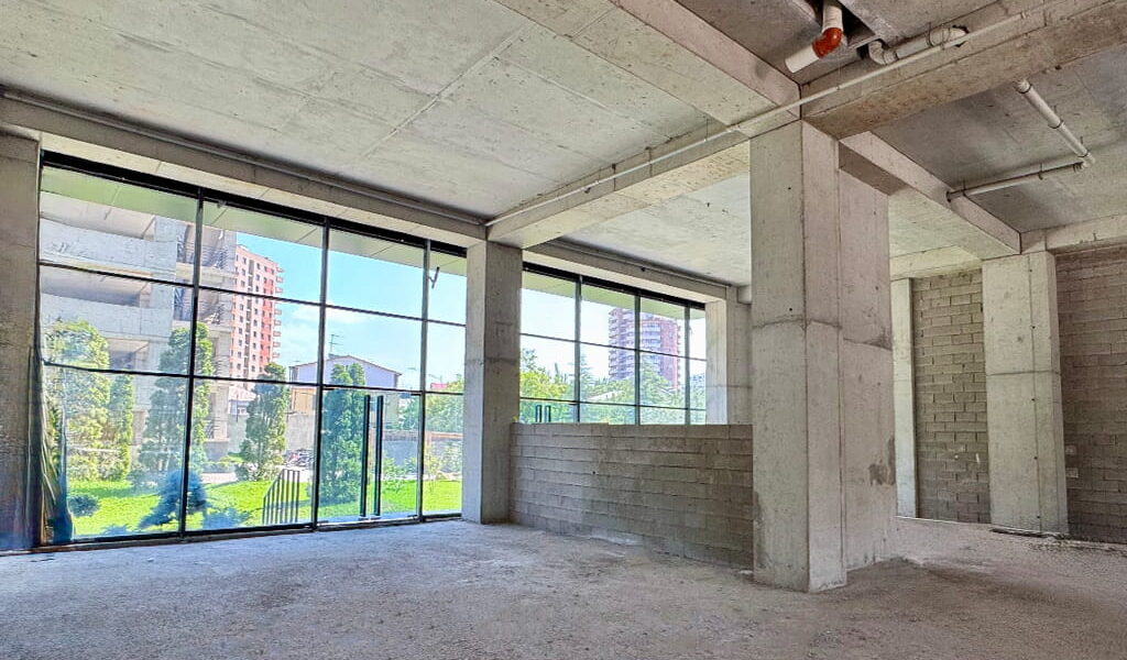 Unfinished concrete interior with large glass windows showing a green outdoor space and buildings outside.