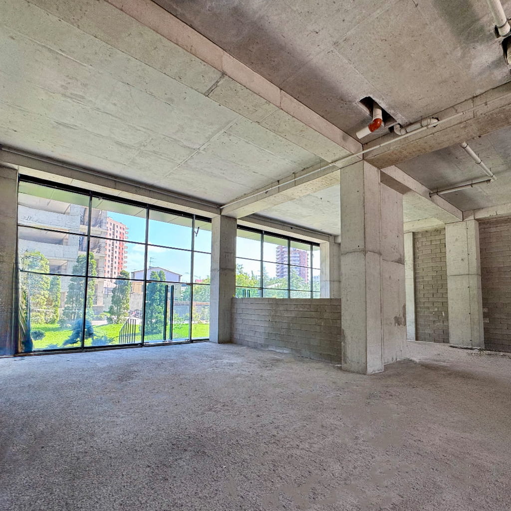 Unfinished concrete interior with large glass windows showing a green outdoor space and buildings outside.