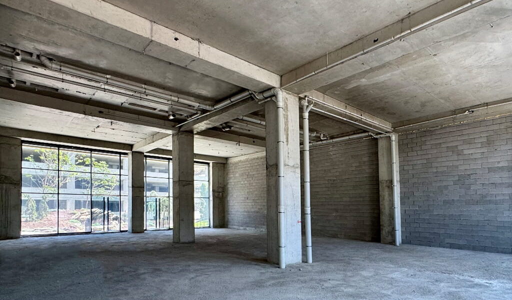 Empty unfinished industrial space with concrete floors, exposed pipes and large back windows.