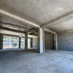 Empty unfinished industrial space with concrete floors, exposed pipes and large back windows.