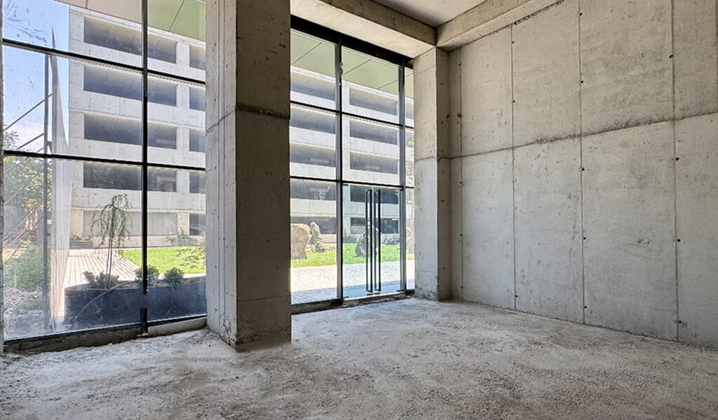 Empty unfinished concrete room with large glass storefront windows and a doorway, daylight outside.