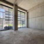 Empty unfinished concrete room with large glass storefront windows and a doorway, daylight outside.