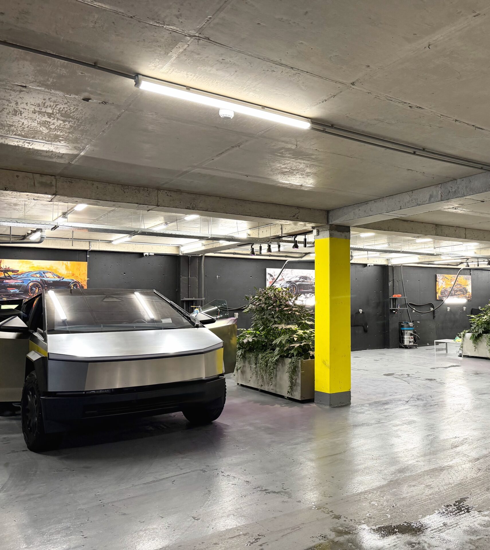 Underground parking garage with exposed concrete ceiling, bright fluorescent lights, and yellow support columns with plants nearby.