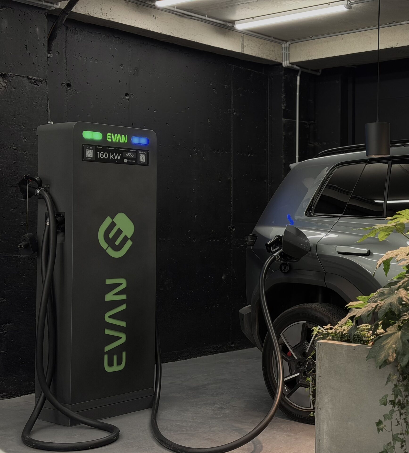 Electric vehicle charging pedestal with green 'EVAN' branding, plugged into a grey SUV in a dark garage.